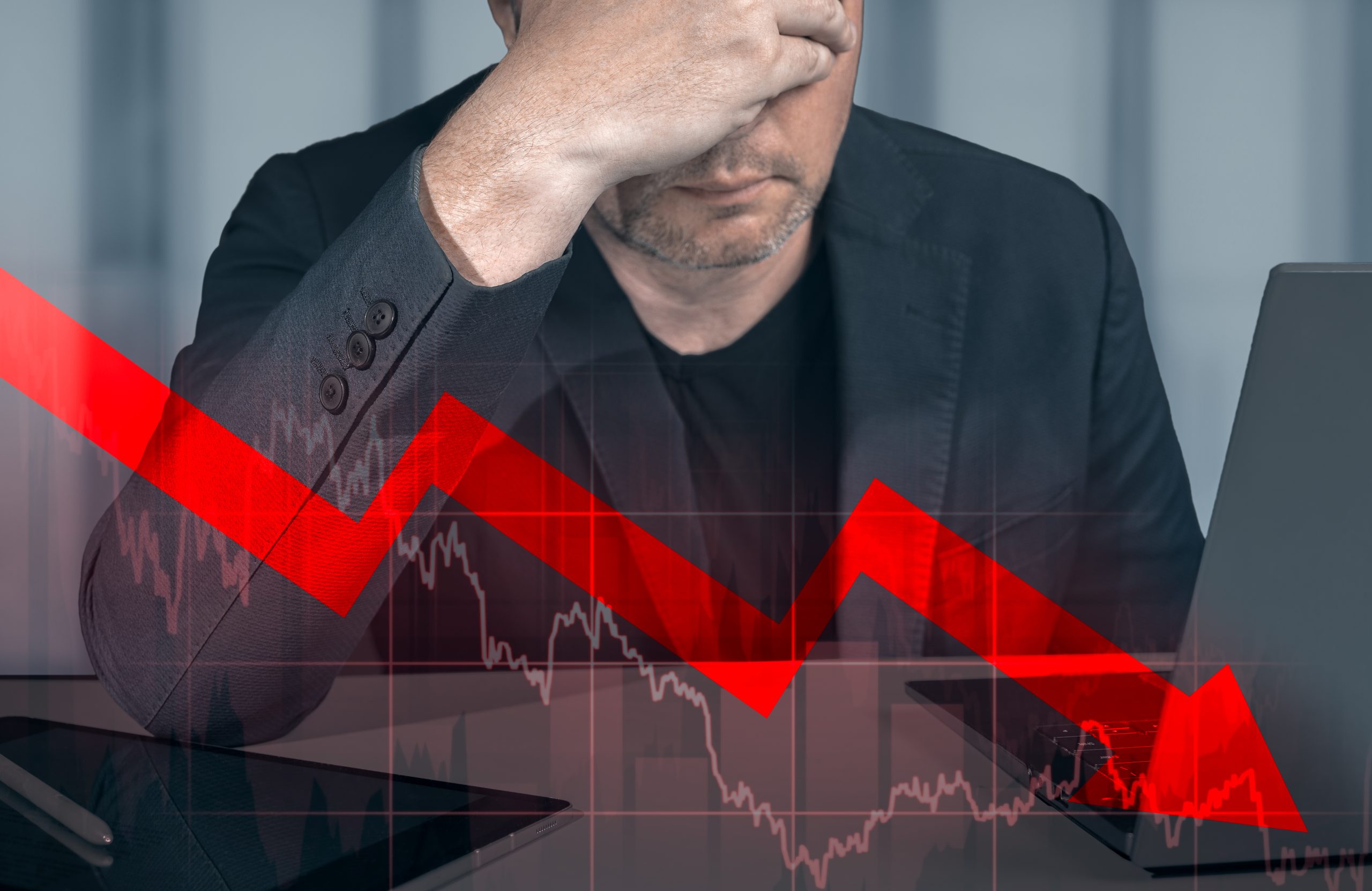 Ein Mann hält sich verzweifelt die Hand vor das Gesicht. Im Vordergrund kann man einen roten sinkenden Pfeil sehen, der eine Finanzkrise darstellen soll.